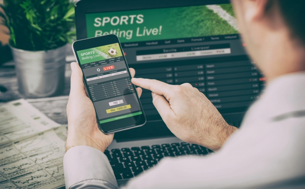 Person using a smartphone to engage with a live sports betting app, with a laptop open nearby showing more betting options.