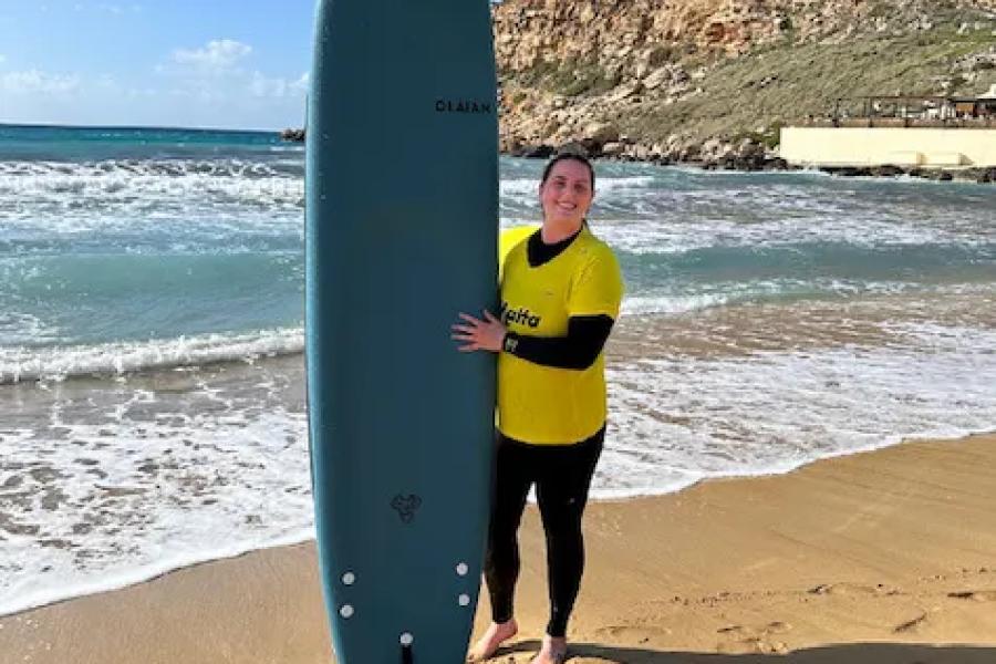 A girl in a yellow shirt and black wetsuit stands on a beach with a blue surfboard.