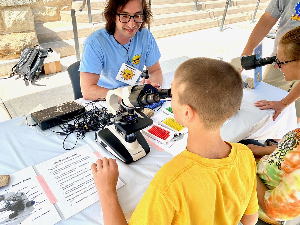 Two young visitors peer into microscopes as a K-POP (Kansas Postdoctoral Outreach Project) team member provides instruction during a recent science event at the museum. Credit: Eleanor Gardner, KU Natural History Museum.
