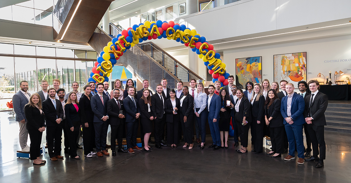 A group of students pose in front of a blue, red and yellow balloon arch in Capitol Federal Hall for the 2024 MBA Big 12 Case Competition