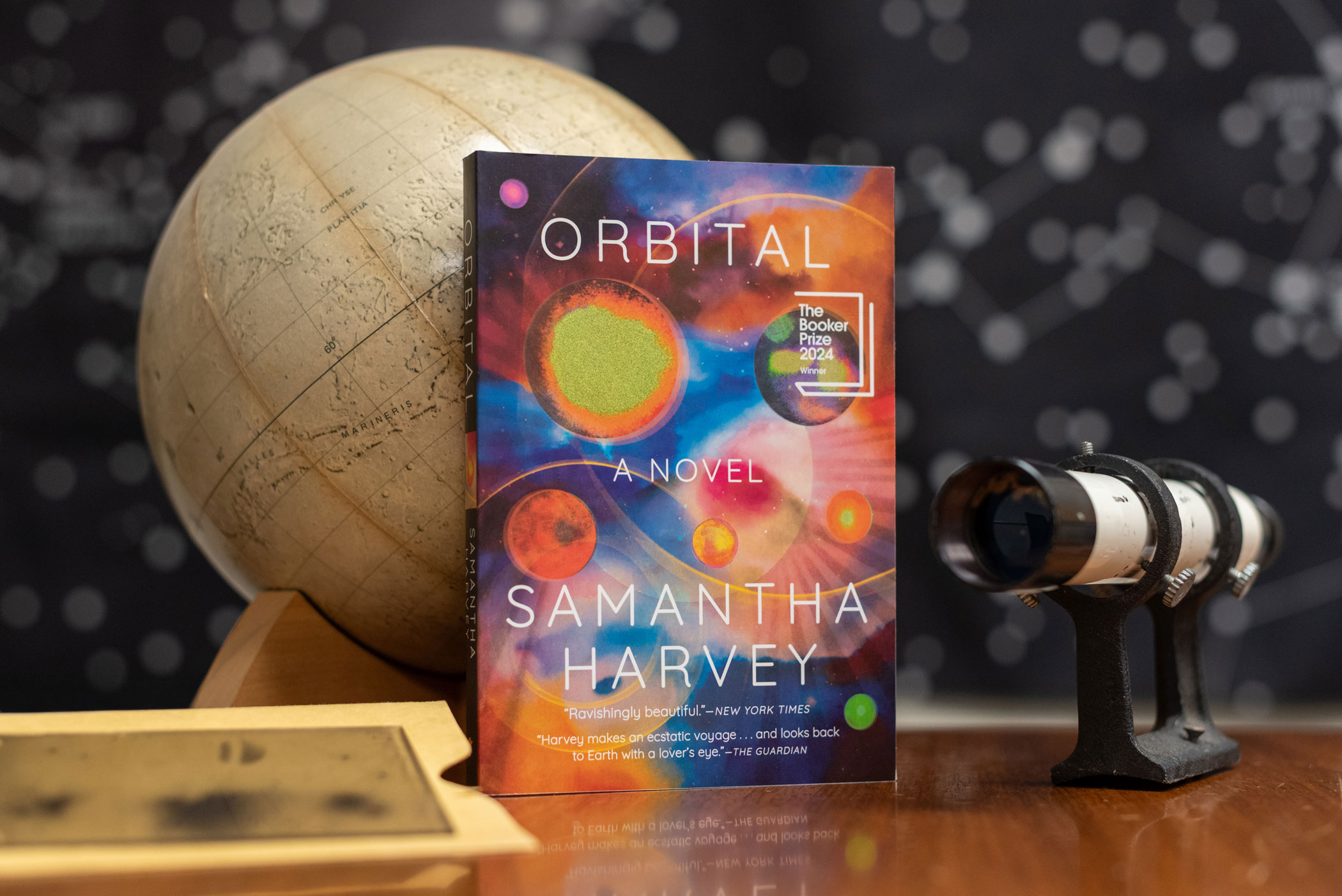 An arranged photo scene featuring the book Orbital among a telescope eye spotter and globe.