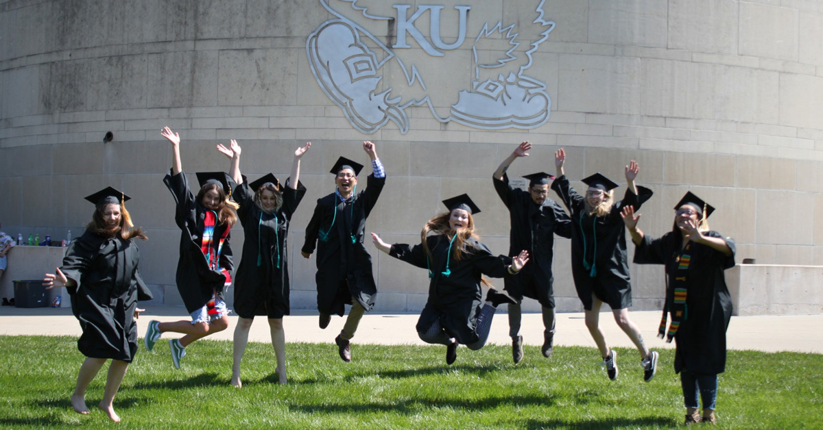 Graduates jump in front of a Jayhawk logo at the KU Edwards Campus