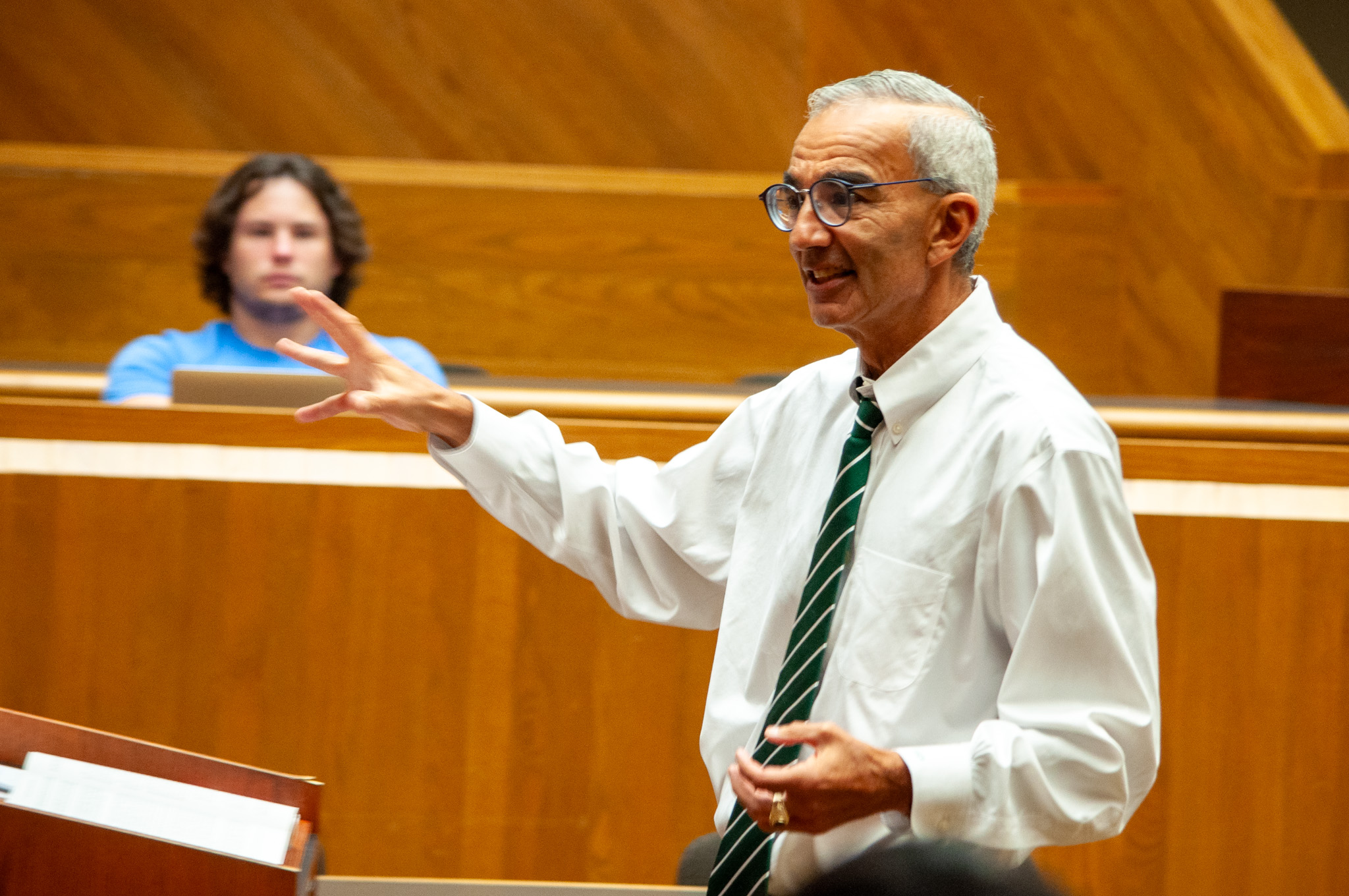 Raj Bhala lectures in front of a white board with a law student in the background.