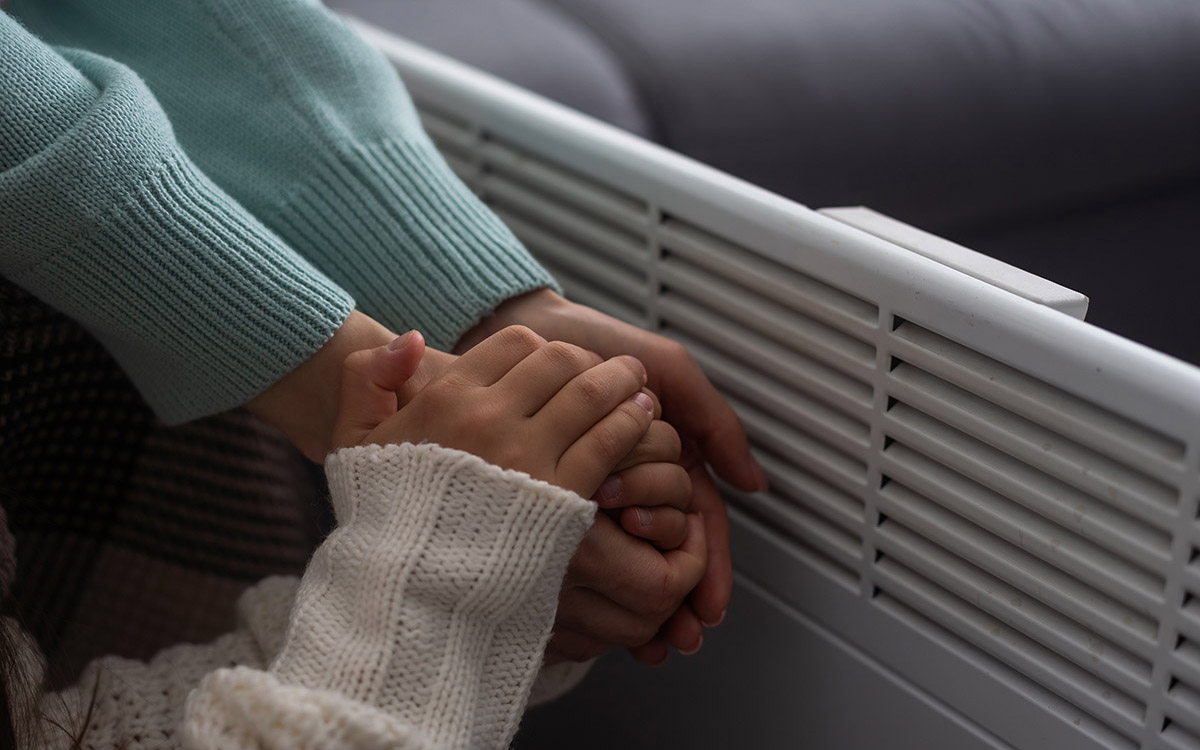 An adult and child hold hands to warm them near a heater.