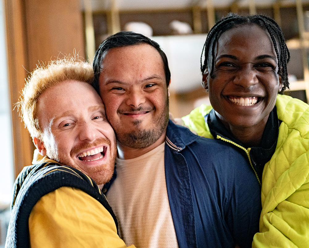 A man with red hair and a beard wearing a yellow shirt and blue vest, a man with dark hair and a beard with a blue jacket who has Down syndrome, and a man with dark hair in braids wearing a bright yellow-green jacket embrace and smile for a photograph together.