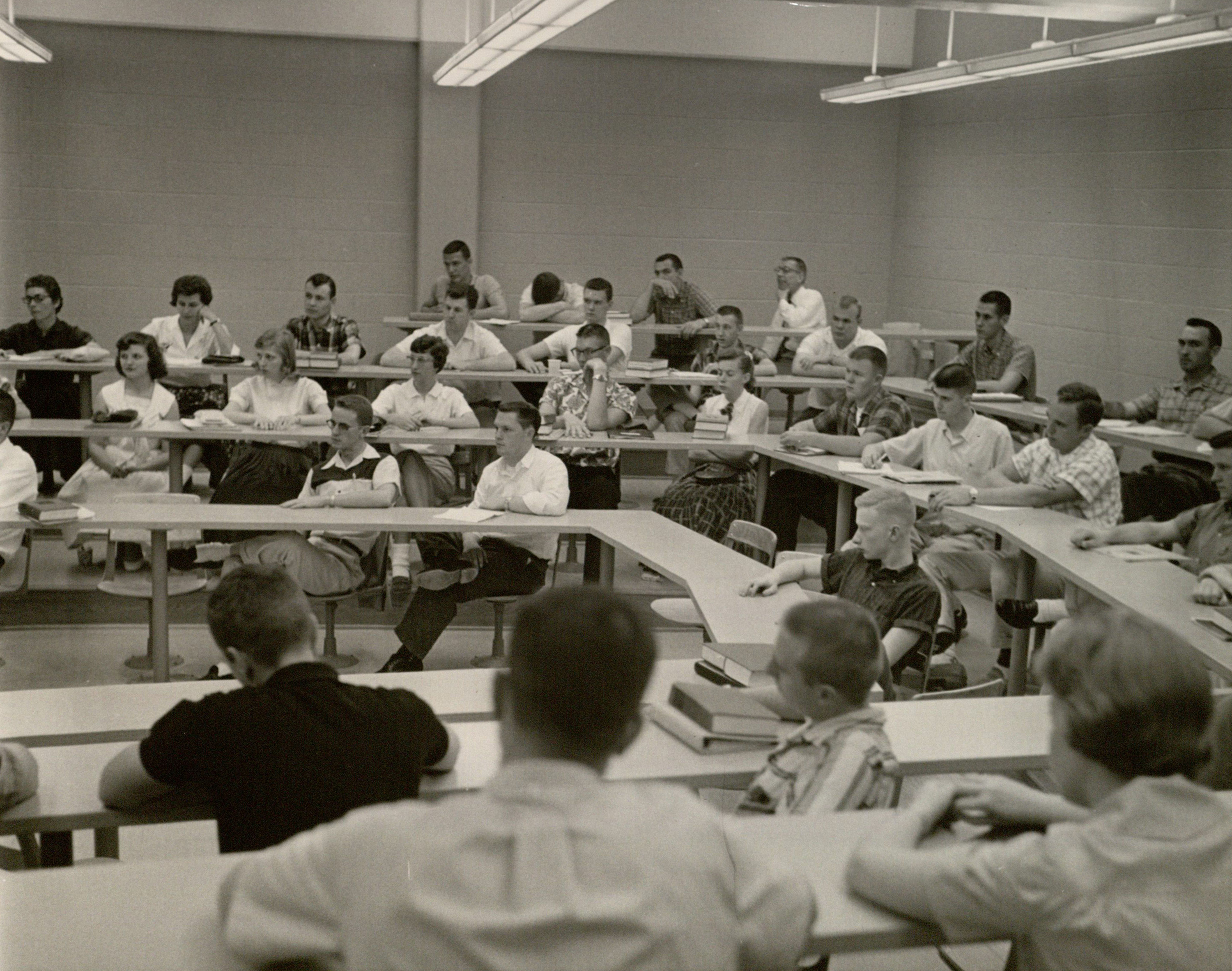 A black and white photo of students in a classroom sitting around tables run along the room.
