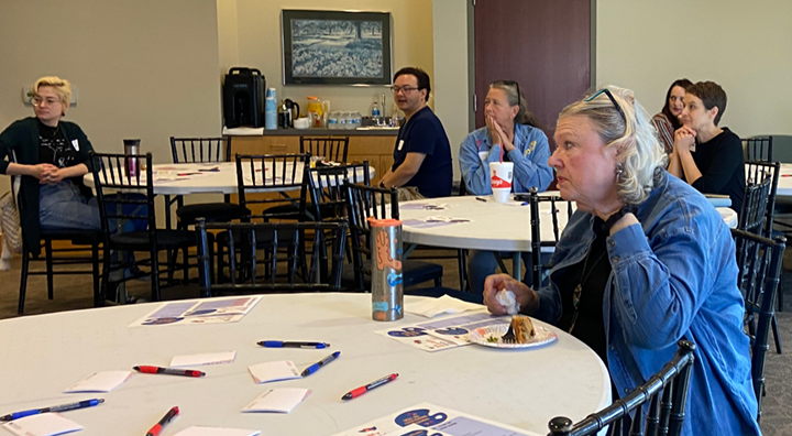Attendees of the November 14 2025 Arts Forum sit around a circular table and watch Nick Carswell present on audio description.