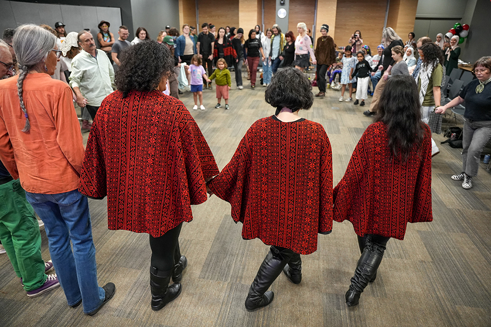 Three women with dark hair wearing matching red garments lead a group dance with adults and children circled around them