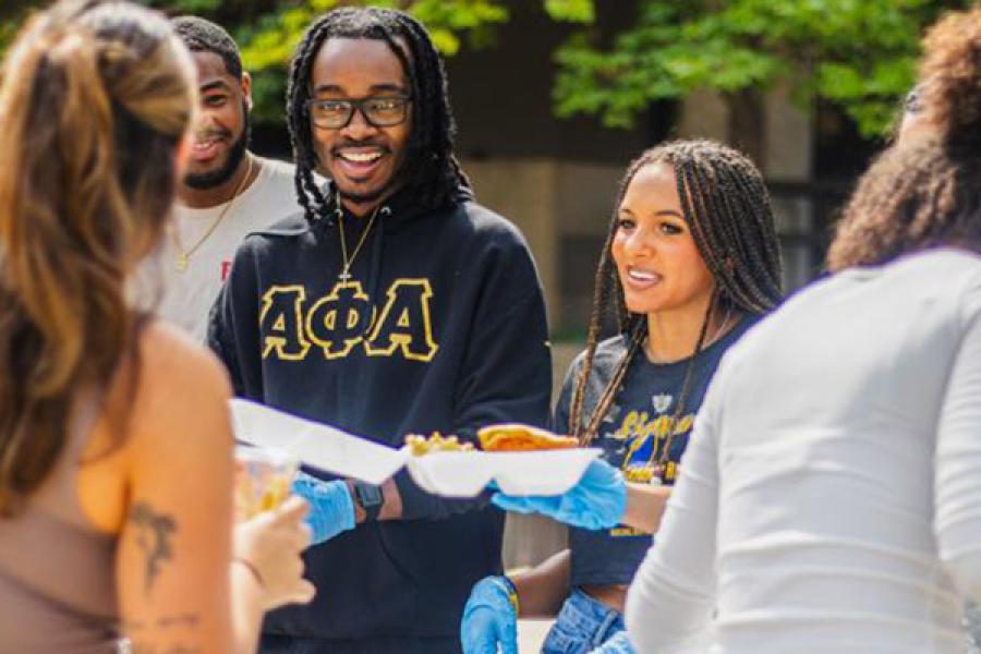 A group of people are gathered outdoors at a food serving event where one individual is wearing a black hoodie with gold Greek letters. Two people are serving food, wearing gloves, and another person is holding a plate.