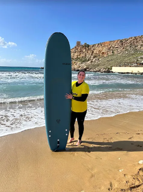 A girl in a yellow shirt and black wetsuit stands on a beach with a blue surfboard.