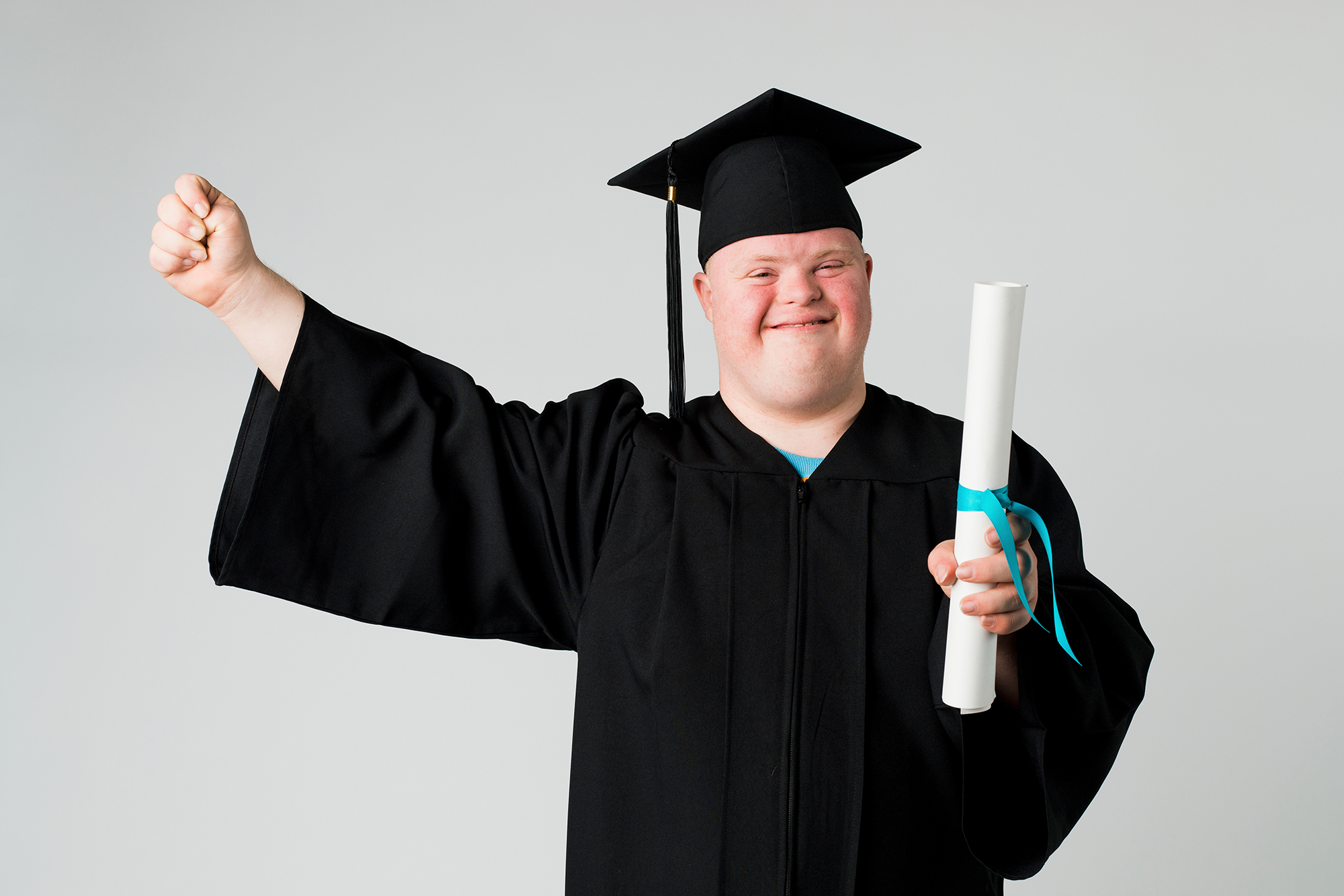 "A man with Down syndrome wearing a graduation outfit smiles and raises a fist in triumph"