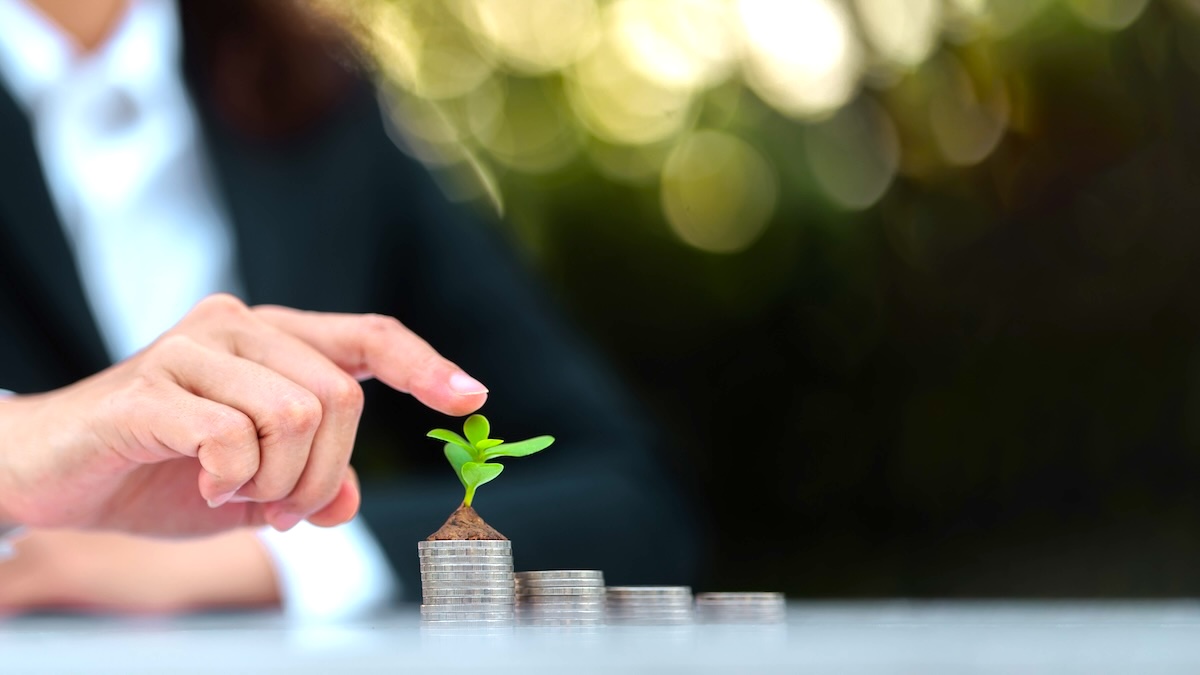 A businessperson touches a green seedling on a stack of growing coins.
