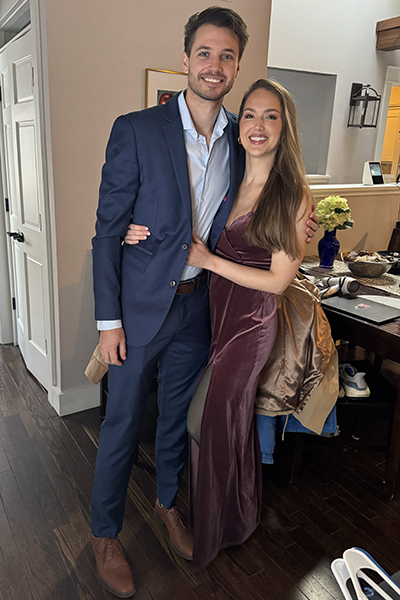 Two individuals in formal attire posing together in a room, with one wearing a blue suit and the other in a purple dress.