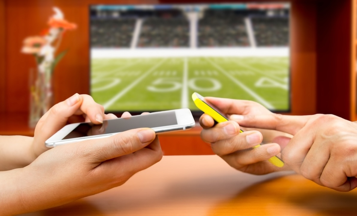 Two people use their phones to bet on a football game with TV in background.