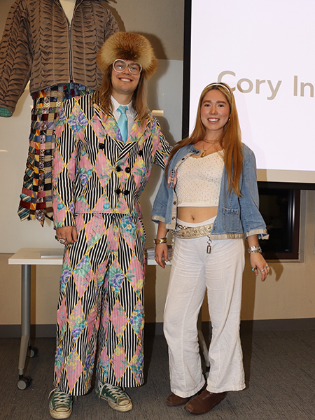 A person wearing a colorful, patterned suit and matching wide-brimmed hat stands next to another person in a denim jacket and light, flowy pants. The background features a clothing display and a partial projection of text on a screen.