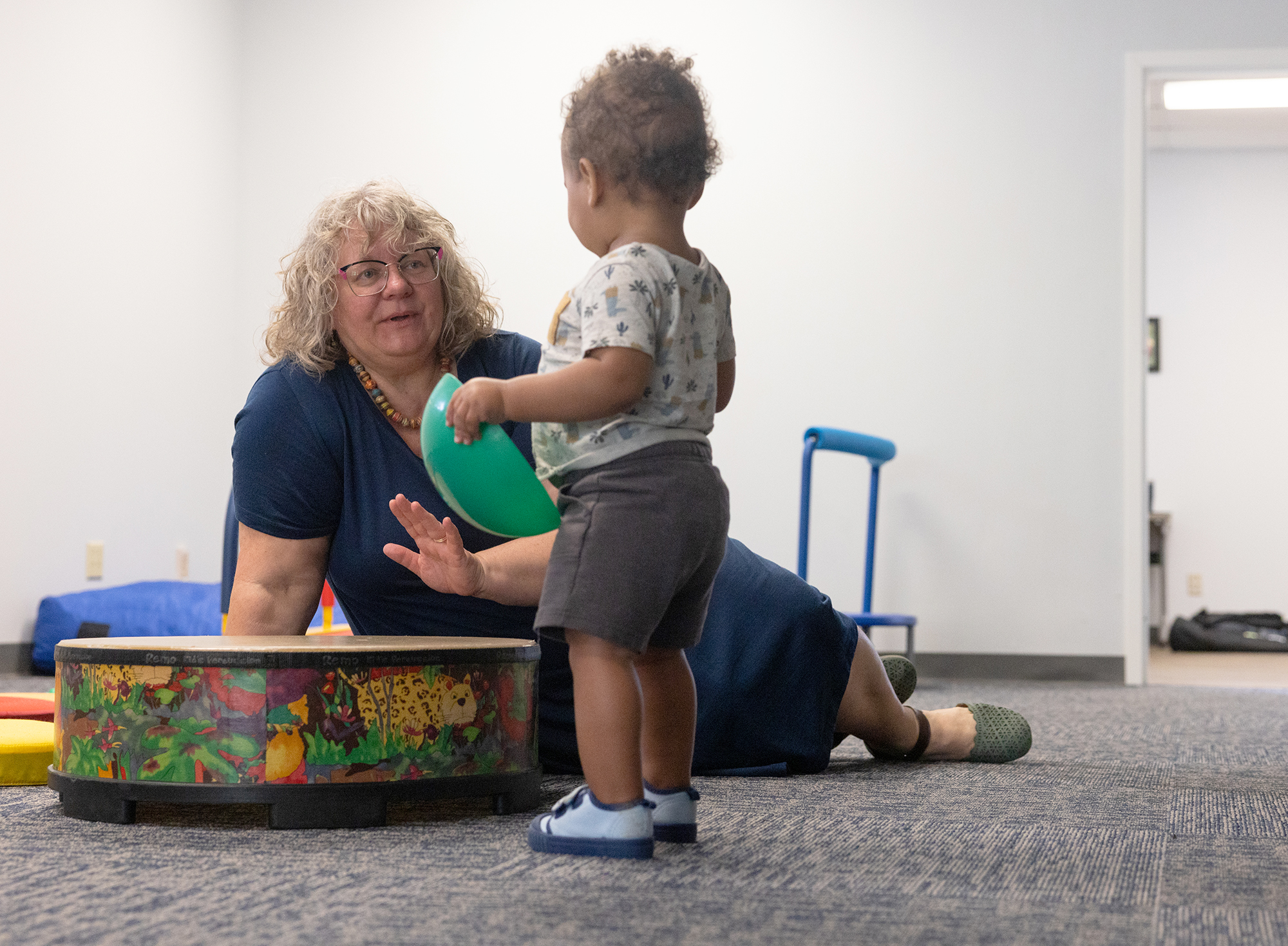 A woman with curly blonde hair sits on the floor playing a drum to a toddler standing next to her.