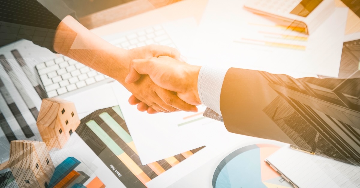 Two unseen people shake hands over a desk littered with business tech and materials.