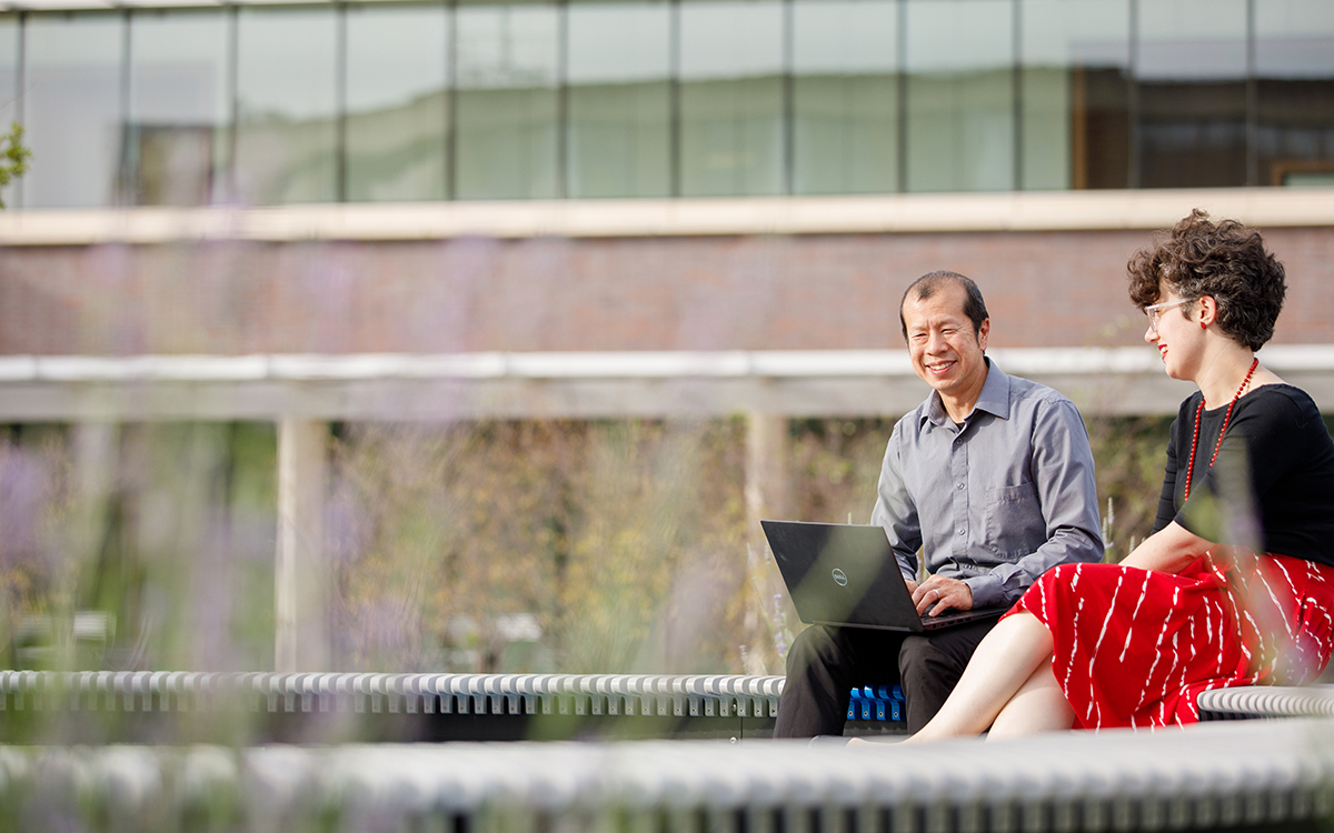 A pair of adult learning sit on a bench with computers outside the BEST Building at the KU Edwards Campus.