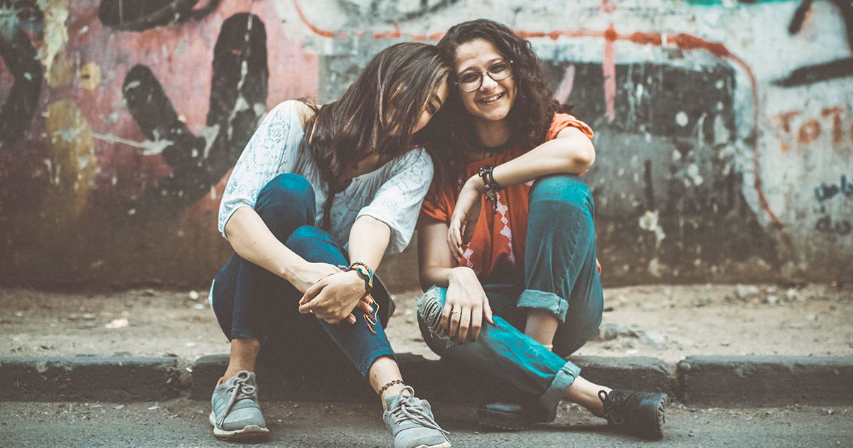 Two young women sitting on sidewalk, smiling and laughing. Stock image.