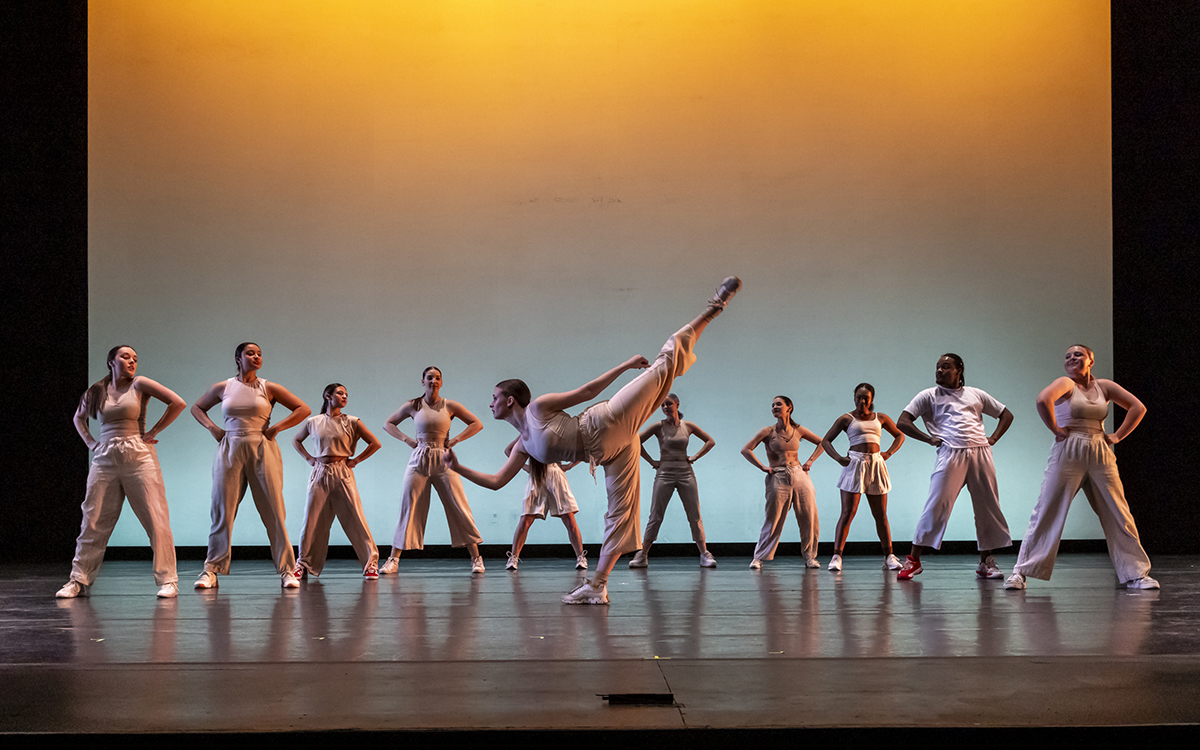 A dancer performing an arabesque onstage with fellow dancers in background.