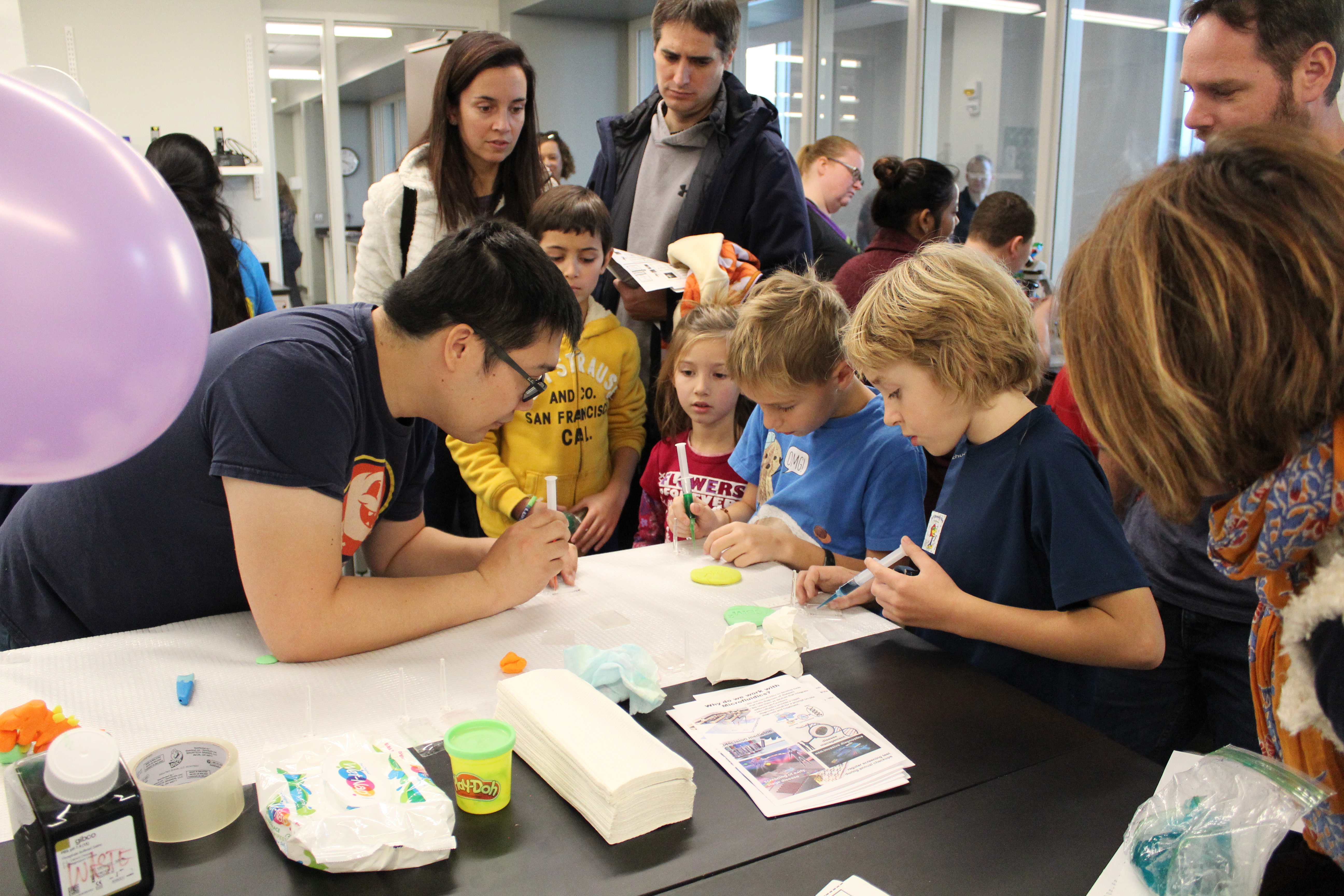 A group of people surrounding a child as they inject color into a glass.