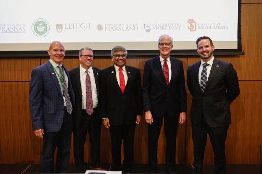 A group of men in suits standing in front of a projector screen with college logos on it