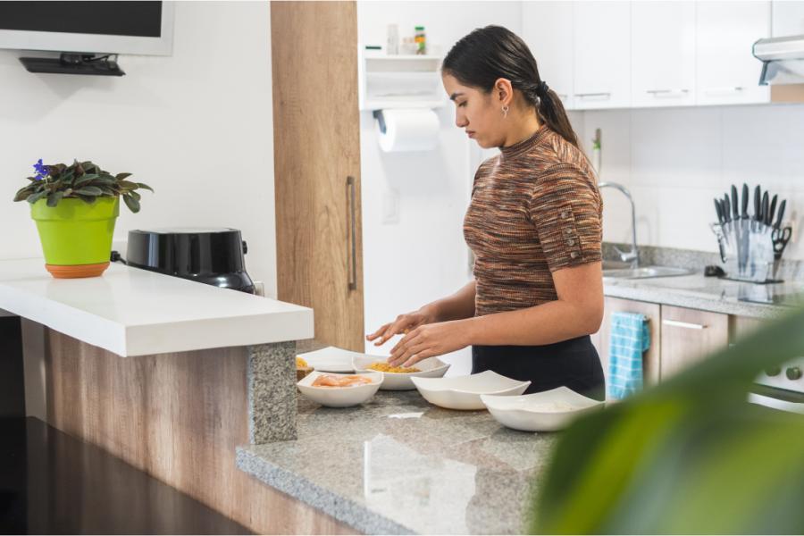 A woman with long dark hair tied back in a ponytail and a brown sweater prepares food in a kitchen