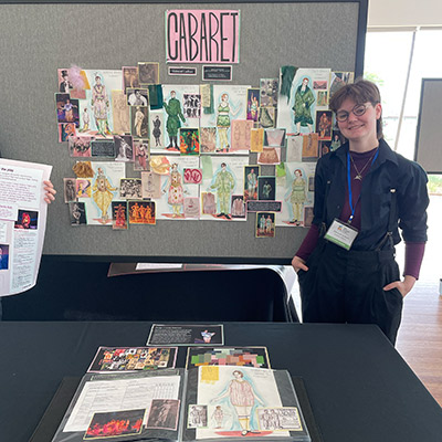 "Student dressed in black with some deep purple accents looks directly into the camera showing an expressive smile. The student’s hands are in their pockets.  The photo shows their booth at a theatre design festival. A table in front and bulletin board in back show their swatches, costume design renderings, and a big sign that reads Cabaret."