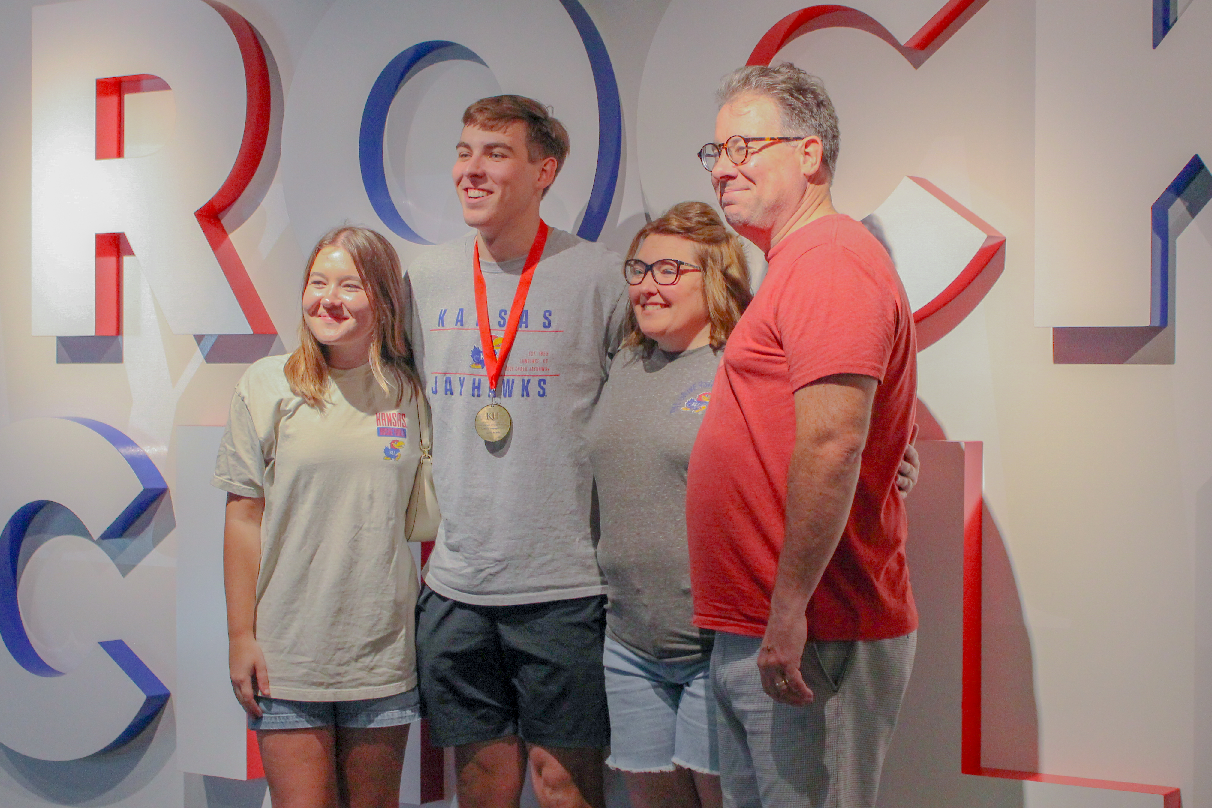 A family posing in front of a "Rock Chalk" sign in the Jayhawk Welcome Center, while a member wears a medal.