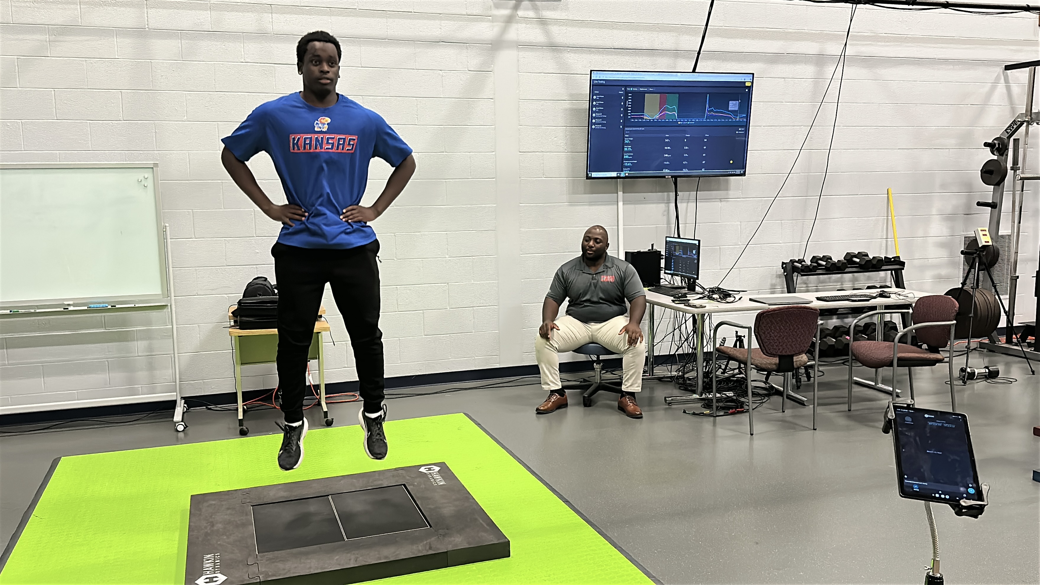 A KU football player jumping vertically on a force plate. Dr. Quincy Johnson sits behind the player with a computer and a screen showing movement data.