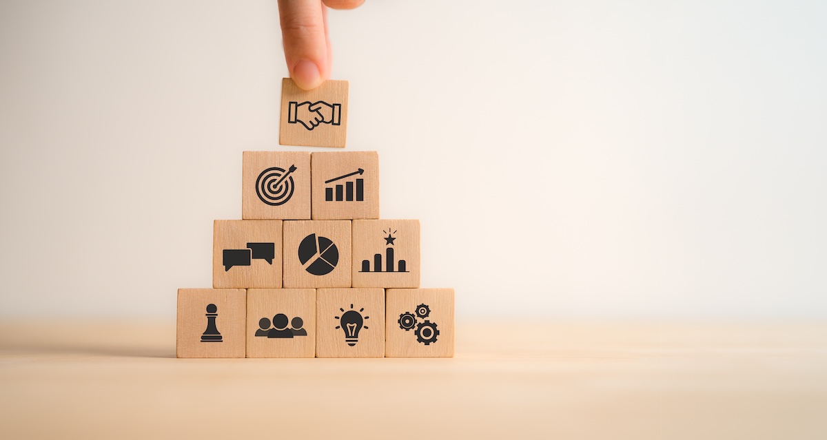 A hand arranges blocks that represent different aspects of business relationships, from ideas to profitability to manufacturing to sales.