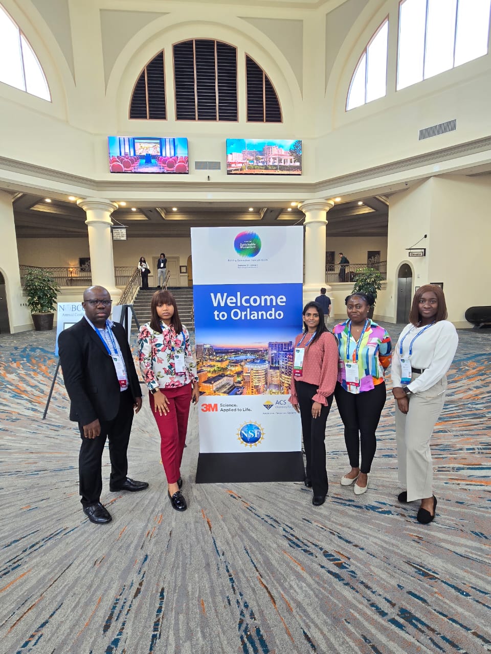 Five KU-NOBCChE members in the lobby of the conference venue, standing on either side of the conference's welcome display.