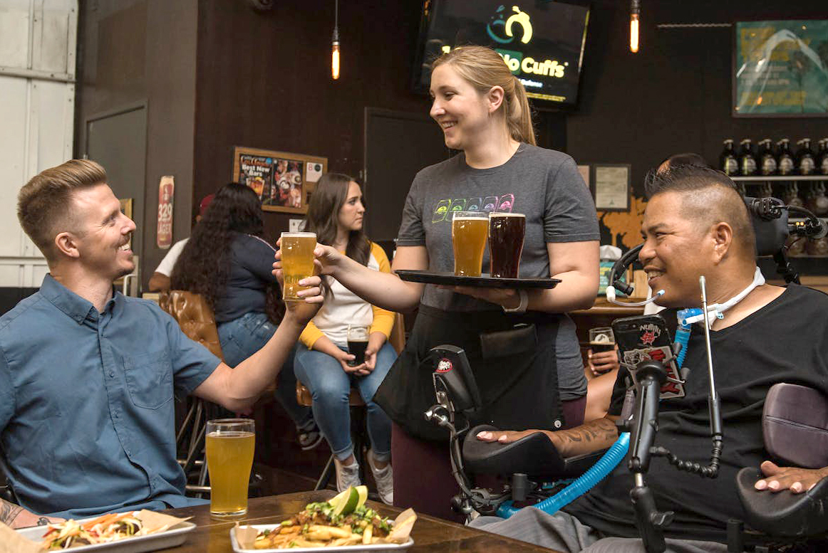 "A man sitting in a motorized wheelchair smiles at his friend who accepts a drink from a server at a restaurant"