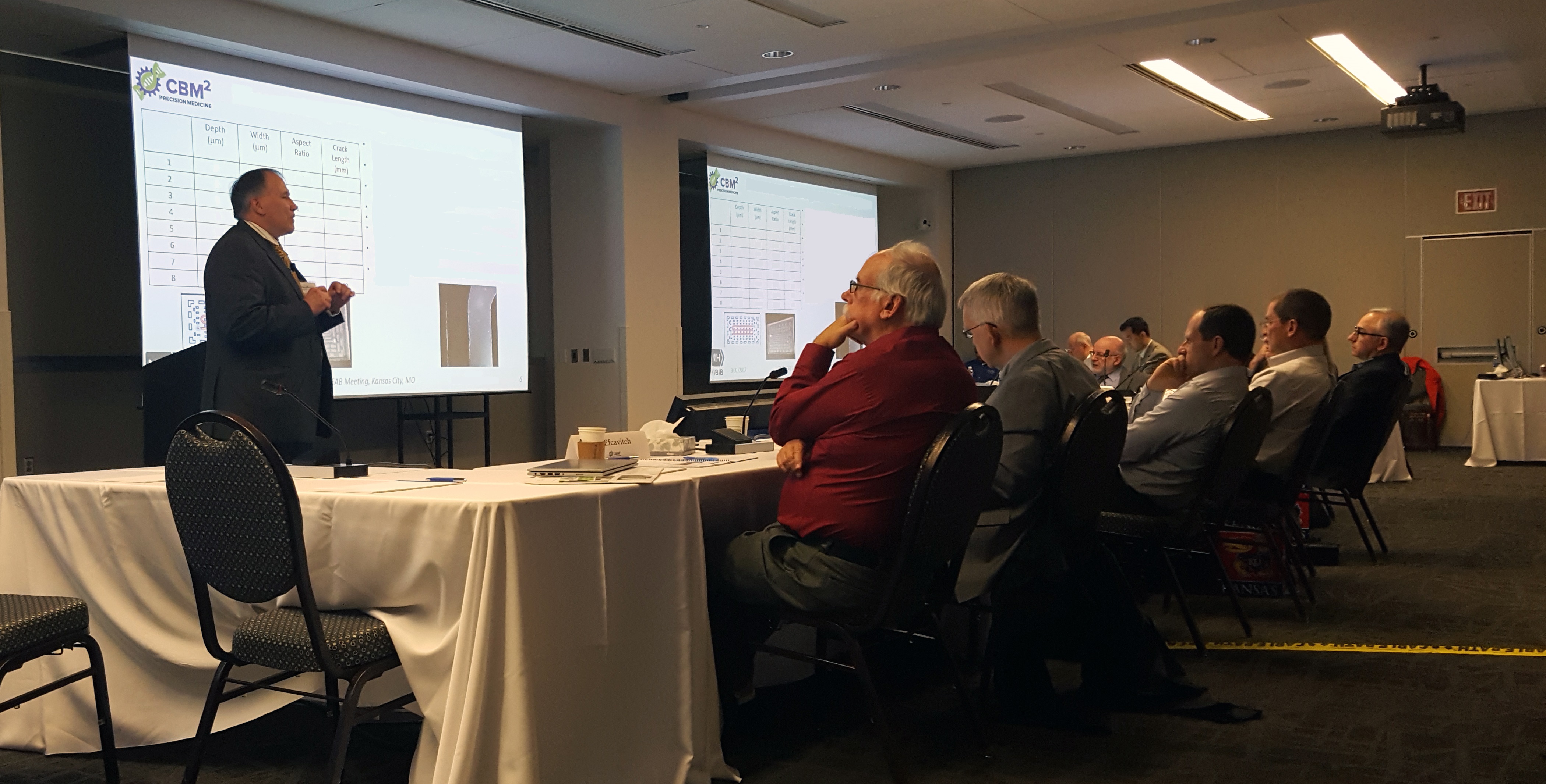 Speaker at the front of a conference room addressing a seated audience at tables.