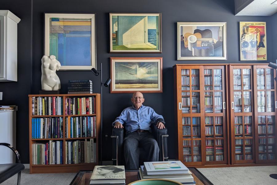 Stephen H. Grabow in his home, seated on a Le Corbusier LC2 Petit Modele Armchair, between two bookshelves in a room decorated with framed artworks and a sculpture.