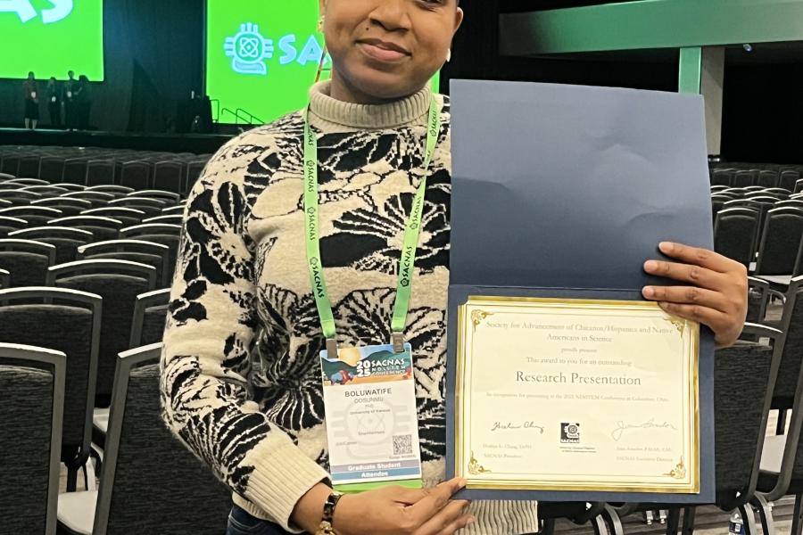Boluwatife holds her Research Presentation award, with the SACNAS conference stage visible in the background