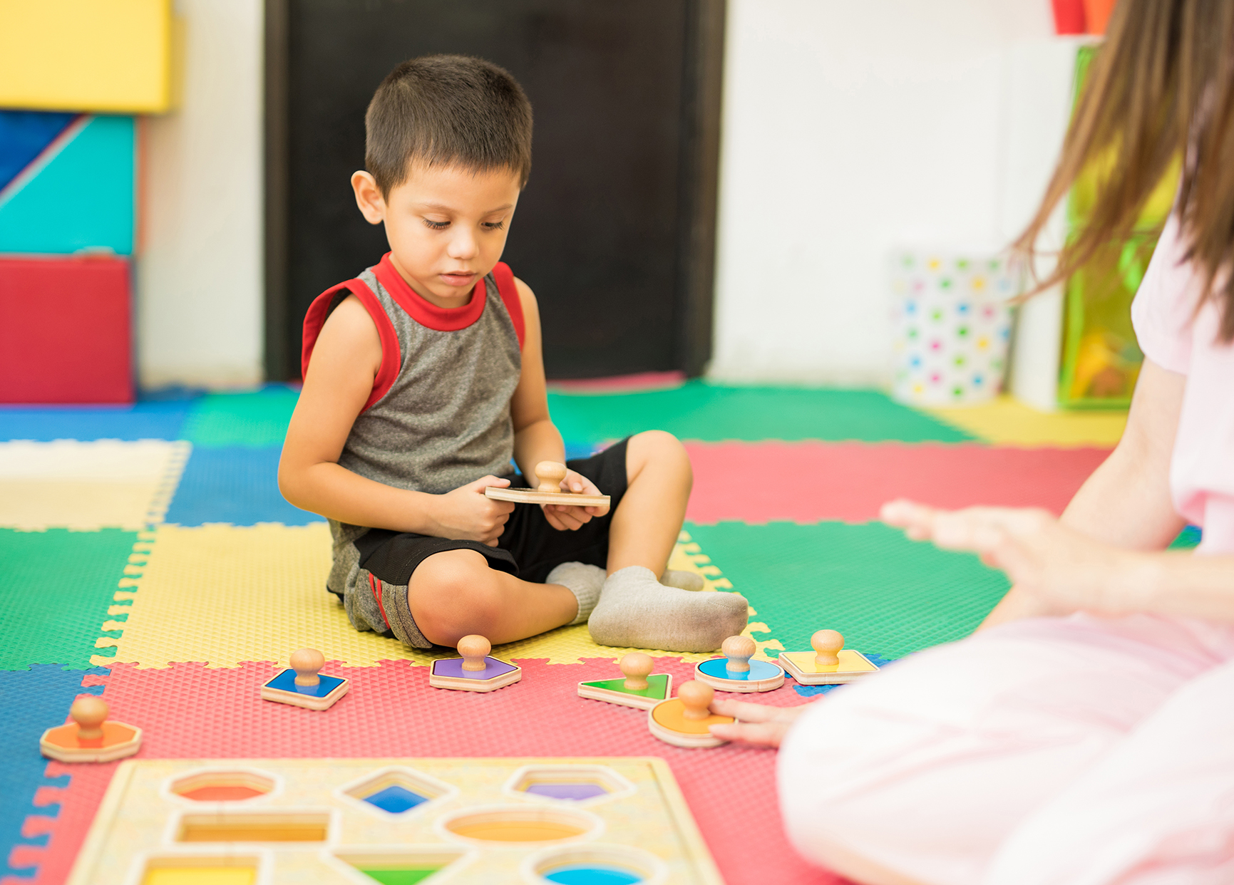 "A young boy plays with a shape puzzle on the floor while an adult sits in front of him"