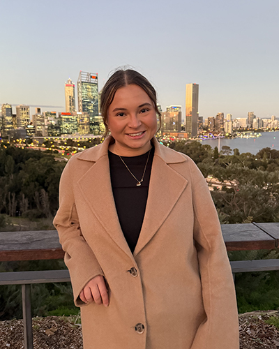 A person in a beige coat smiling at the camera with the Perth skyline illuminated by sunset in the background.