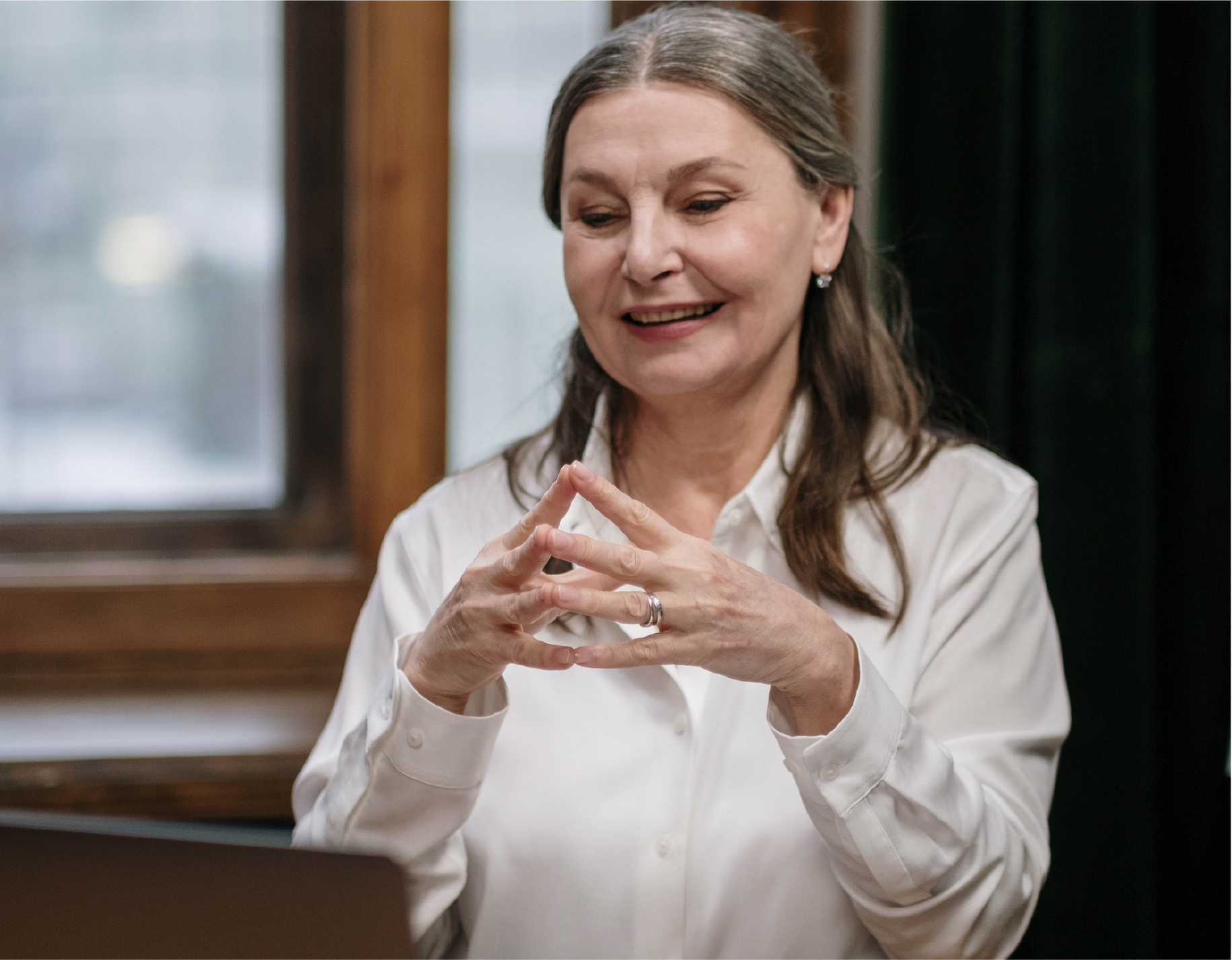 "A woman with long gray hair wearing a white blouse smiles while looking at a laptop"