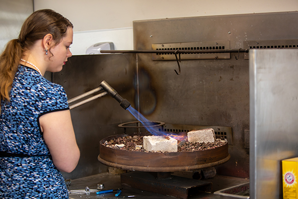 A person is using a blowtorch to heat materials placed in a large metal pan filled with rocks. The setting appears to be a workshop or kitchen area with metal surfaces.