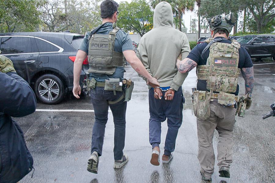 A photograph of ICE agents detaining a man and walking away from the camera with their backs turned as a man is handcuffed behind his back.