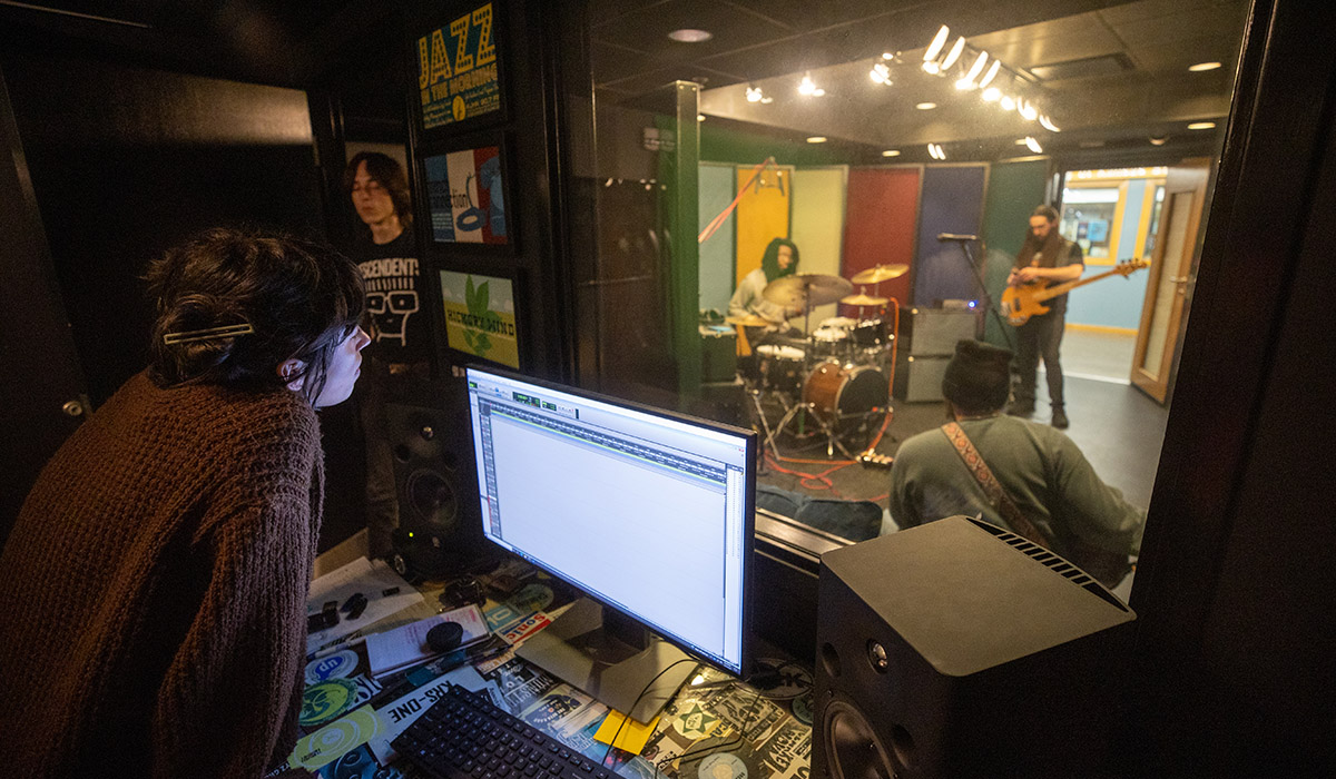 Two students in front of audio equipment board watching musicians perform in closed glass recording studio.