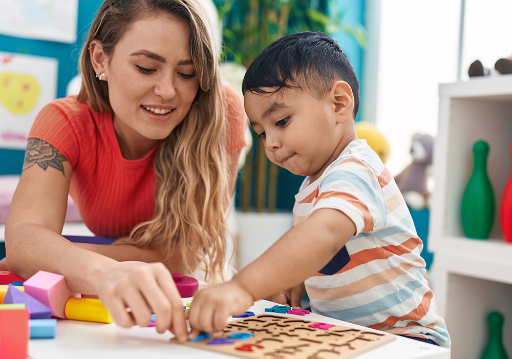 A woman with long light brown hair in a red shirt plays with a toddler with dark hair and a striped shirt as the put together an alphabet puzzle.