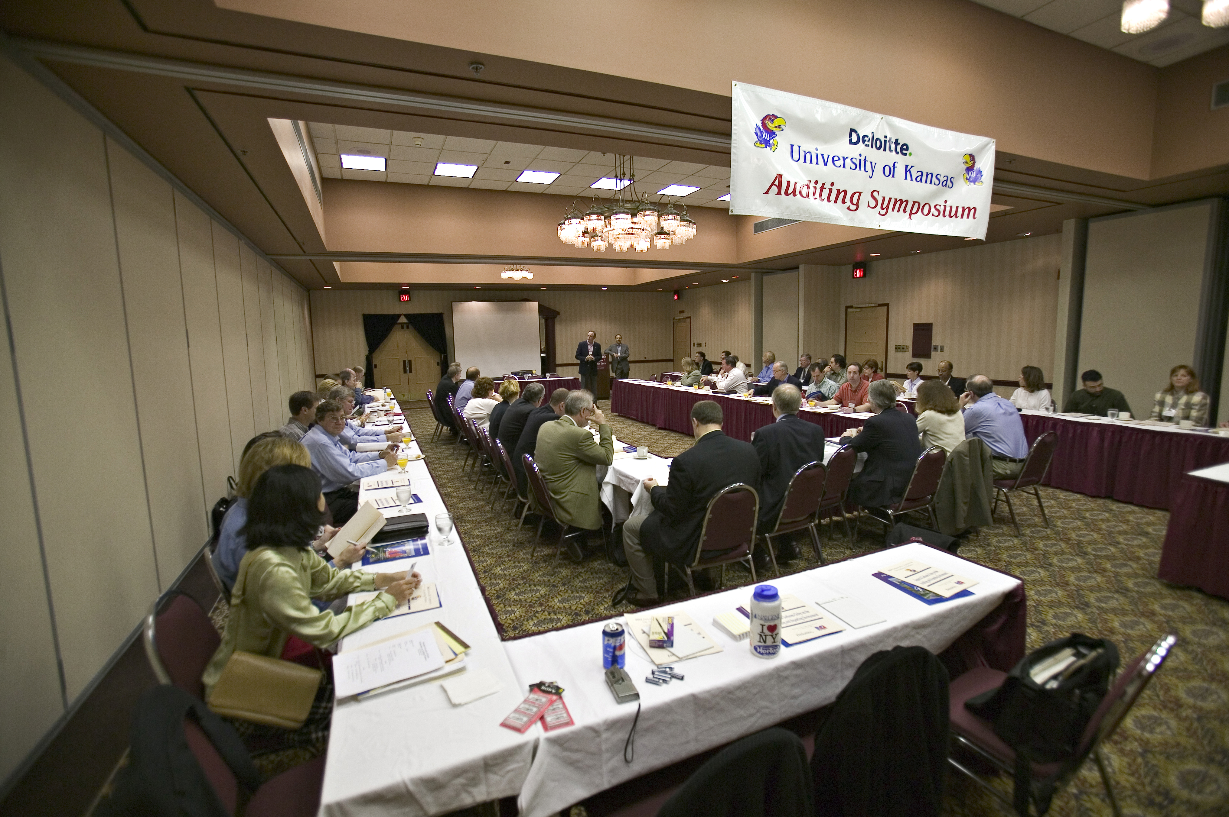 Attendees seated at tables listening to a speaker at the Deloitte/KU Auditing Symposium in a conference hall.