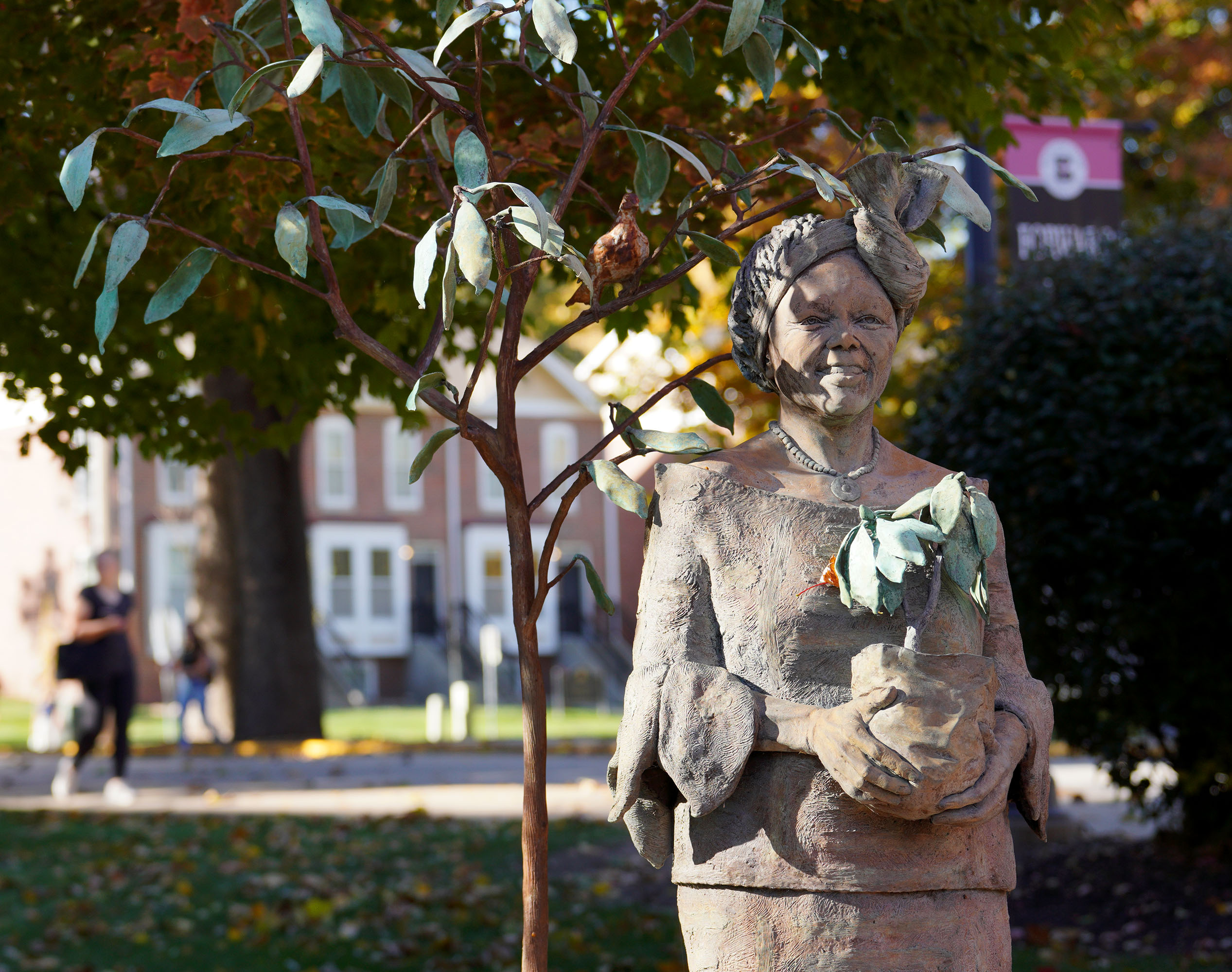 A bronze statue of Wangari Maathai surrounded by fall foliage; beside her is a small tree, and in her hands, a bright green sapling.