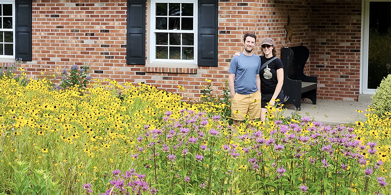 two people standing in front of a brick house with wildflowers covering front yard.