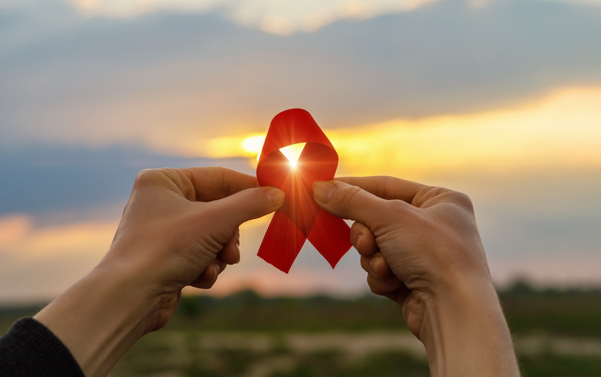 Two hands hold up an AIDS ribbon to the sky, with the sun shining through the top loop.