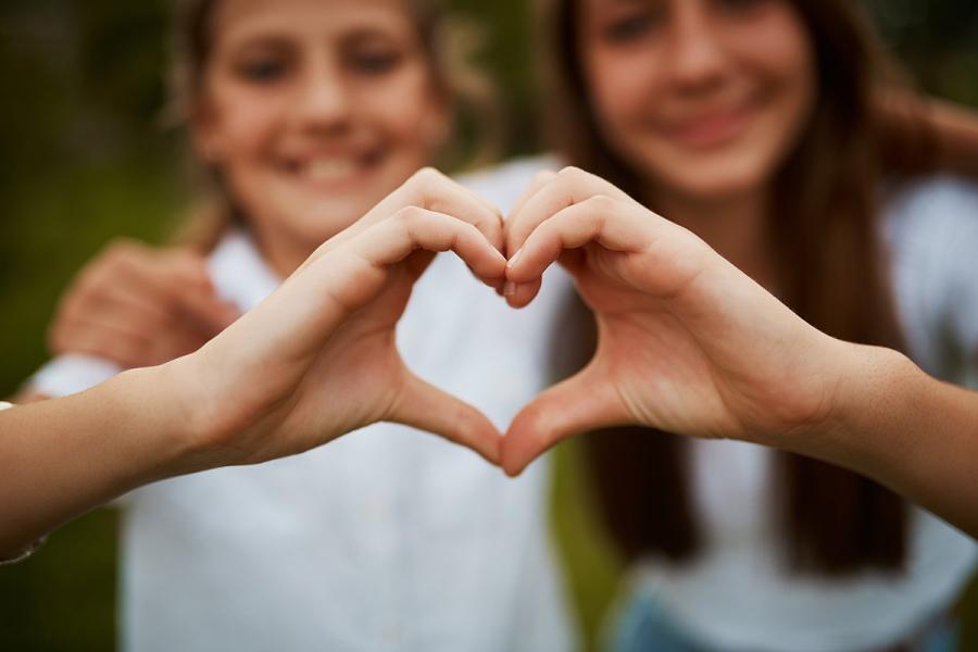 A photo of two young women holding their hands together to form a heart in the foreground as their faces are out of focus in the background.