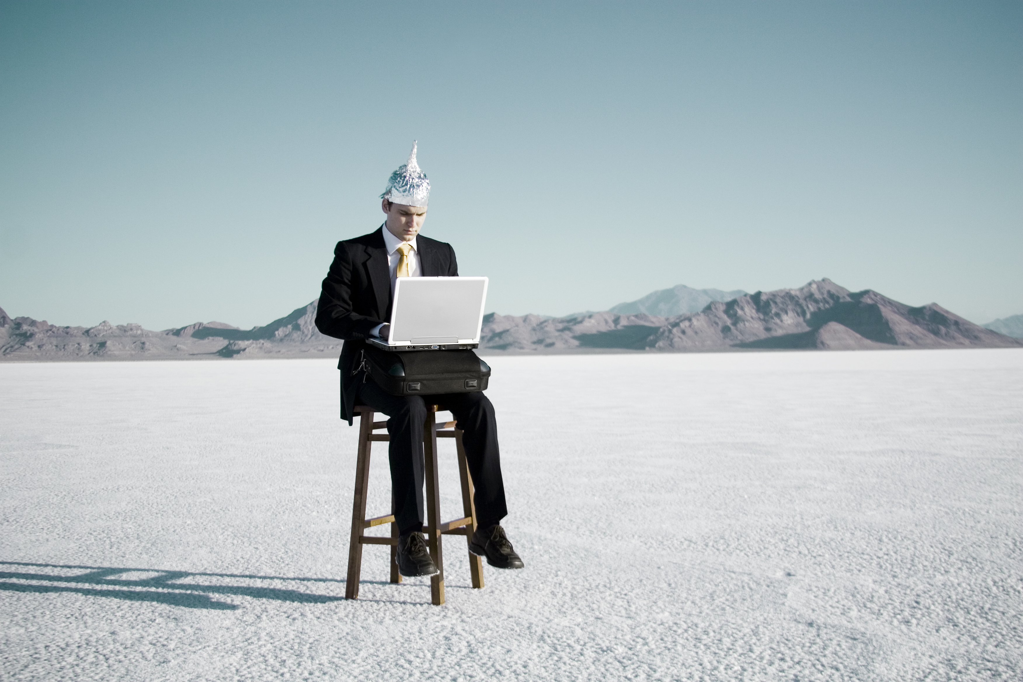 Man in a suit wears a tinfoil hat while sitting on a stool in the desert and working on his laptop