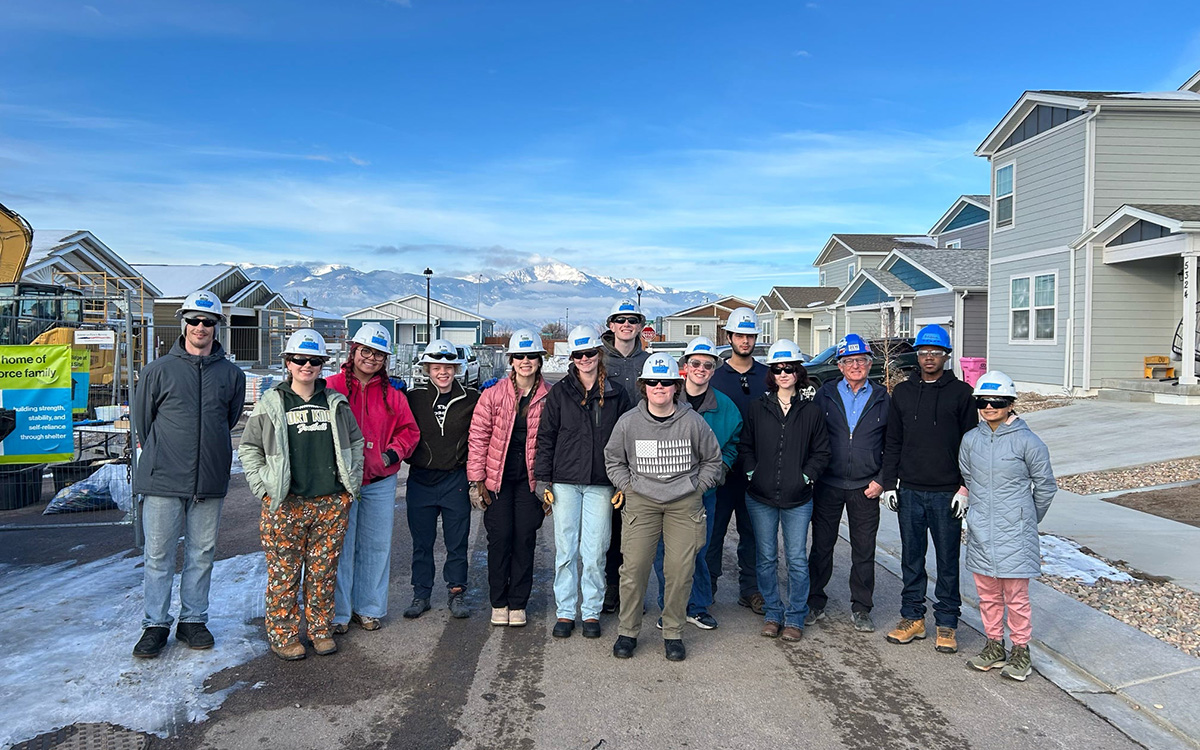 Group of smiling students wearing hard hats and winter jackets, standing with homes and mountains in the background.